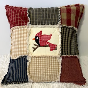 May include: A square rag quilt pillow featuring a nine-patch design with frayed seams. The center square displays an appliqued red cardinal bird perched on a brown branch against a cream background. Surrounding squares include red, black, and tan plaid and gingham patterns.