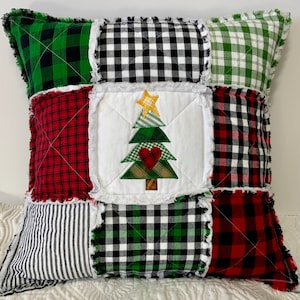 May include: A patchwork Christmas pillow with a central white square featuring an embroidered Christmas tree with a star and heart. The pillow is composed of various plaid and striped fabric squares in red, green, black, and white.