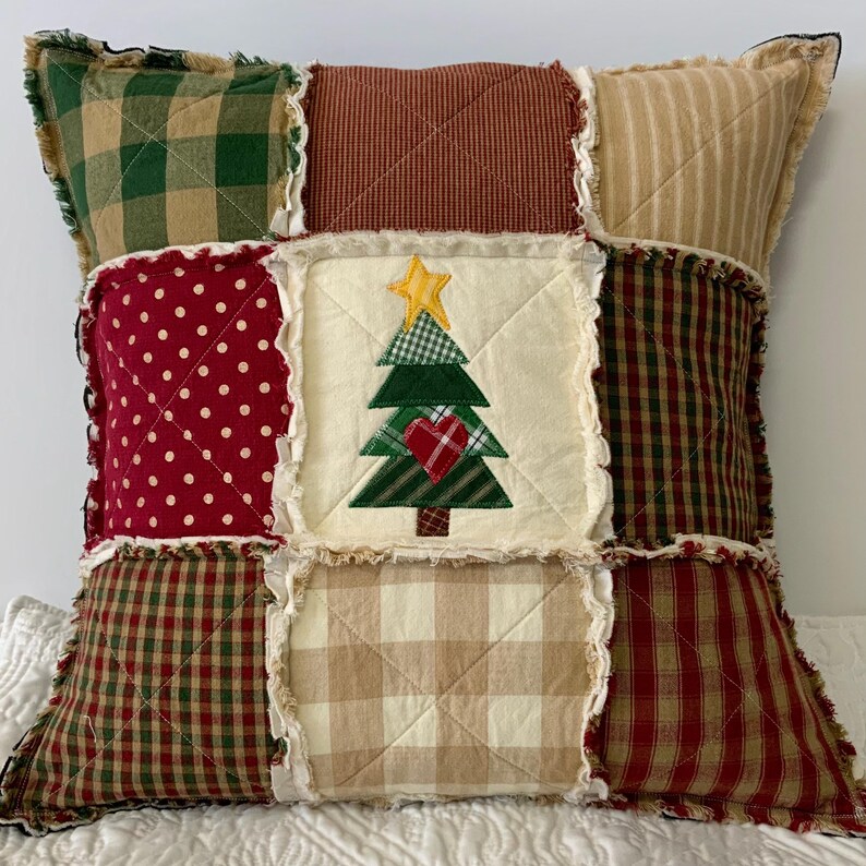 May include: A patchwork pillow with a Christmas tree design in the center. The pillow is made of various fabrics in shades of red, green, brown, and white. The tree is green with a yellow star on top and a red heart in the center.