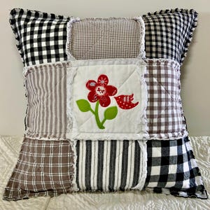 May include: A patchwork decorative pillow with a floral design. The pillow features a central white square with a red flower and bird applique. The surrounding patches are various patterns in black, white, and brown, including gingham, stripes, and plaid.