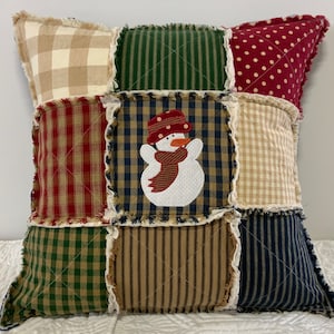 May include: A patchwork pillow with a snowman applique. The pillow features various fabrics in red, green, tan, and navy blue, with a fringed edge. The snowman has a red hat and scarf.