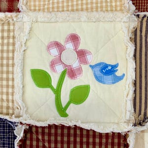 Peut inclure: Un carr&eacute; de tissu blanc avec une fleur &agrave; carreaux rose et blanc, des feuilles vertes et un oiseau bleu en appliqu&eacute;. Le tissu est entour&eacute; d'une bordure blanche effiloch&eacute;e.