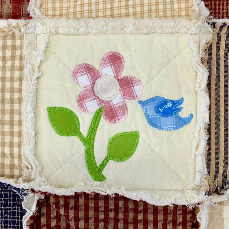 Peut inclure: Un carr&eacute; de tissu blanc avec une fleur &agrave; carreaux rose et blanc, des feuilles vertes et un oiseau bleu en appliqu&eacute;. Le tissu est entour&eacute; d'une bordure blanche effiloch&eacute;e.