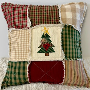 May include: A patchwork pillow with a Christmas tree design in the center. The pillow is made of various plaid fabrics in shades of red, green, and beige. The tree is appliquéd with a red heart and a yellow star.
