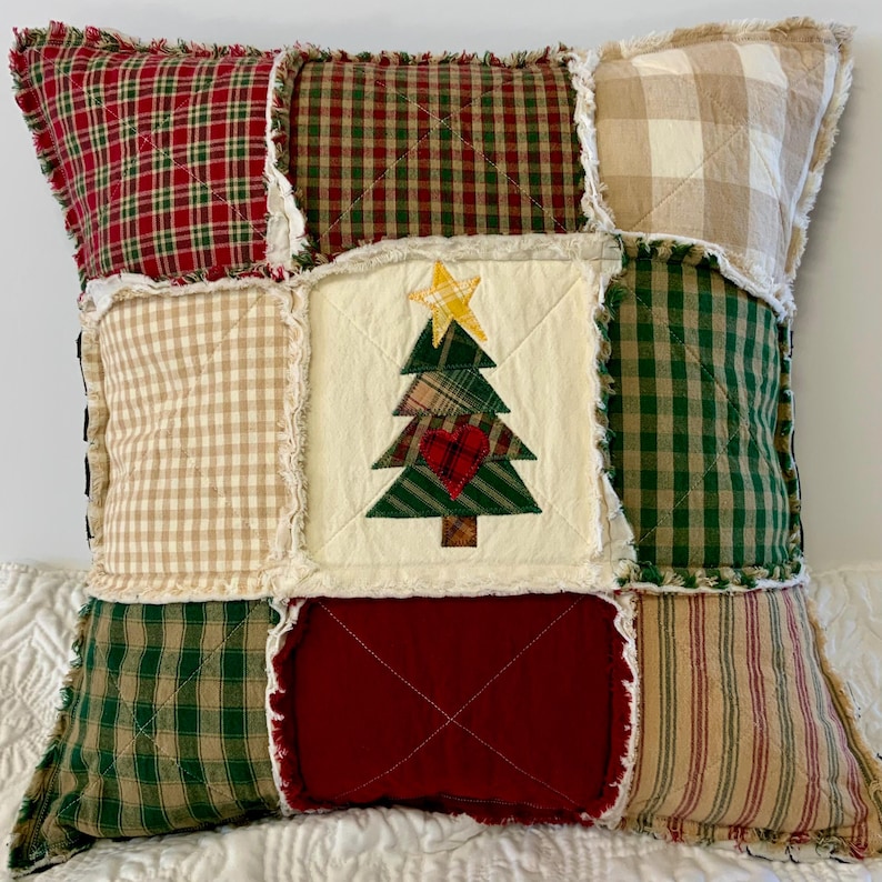 May include: A patchwork pillow with a Christmas tree design in the center. The pillow is made of various plaid fabrics in shades of red, green, and beige. The tree is appliquéd with a red heart and a yellow star.