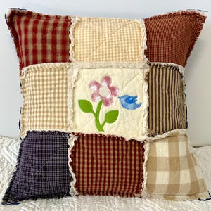 Peut inclure: Un coussin patchwork avec un motif floral et une applique d'oiseau bleu. Le coussin est fait de diff&eacute;rents tissus dans des tons de rouge, marron, beige et bleu. Le coussin a un bord effiloch&eacute;.