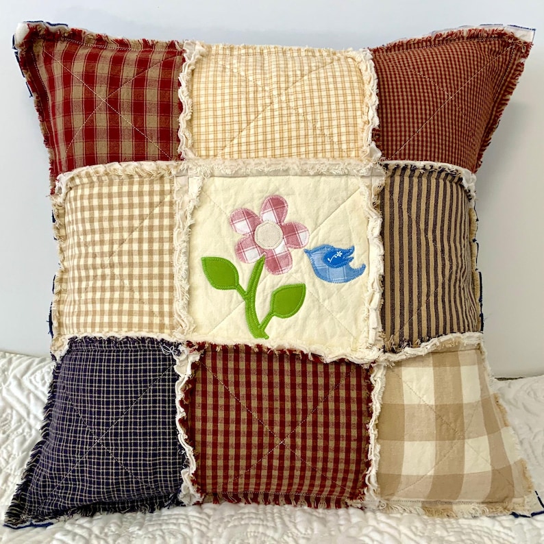 Peut inclure: Un coussin patchwork avec un motif floral et une applique d'oiseau bleu. Le coussin est fait de diff&eacute;rents tissus dans des tons de rouge, marron, beige et bleu. Le coussin a un bord effiloch&eacute;.