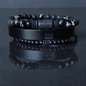 Peut inclure: Trois bracelets noirs avec différentes textures et designs. Le bracelet supérieur présente des perles mouchetées noires et blanches. Le bracelet du milieu a une bande de cuir avec le texte "IRON WOLF". Le bracelet inférieur a de petites perles rondes.