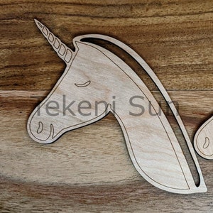 May include: A wooden cutout of a unicorn head with a closed eye and a horn with a spiral design. The cutout is on a woodgrain background.