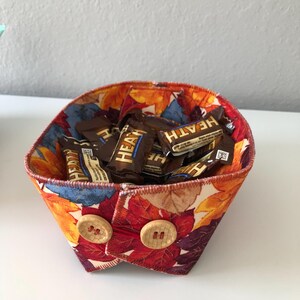 May include: A fabric basket with a fall leaf print in shades of orange, red, and yellow. The basket is filled with individually wrapped chocolate candy bars. The basket has two wooden buttons on the front.