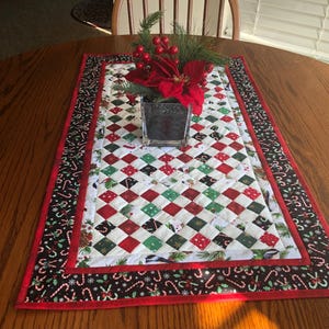 May include: A Christmas-themed table runner with a quilted design. The center features a pattern of red, green, and white diamond shapes. The border is black with candy cane and snowflake motifs, framed by a red edge. A floral arrangement sits on top.