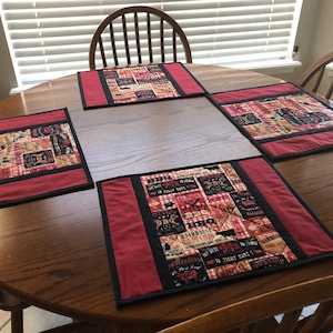 May include: Four red and black placemats with a patchwork design featuring a BBQ theme. The placemats have a black border and red trim. The placemats are on a wooden table.
