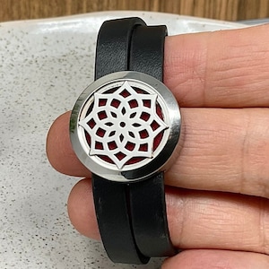 May include: A black leather bracelet with a silver diffuser charm featuring a mandala design. The bracelet is shown with a small bottle of essential oil and three red felt pads.