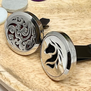 May include: Two silver car air fresheners with decorative designs. One features a floral pattern with a red centre, the other has a horse head design. Both are on a wooden surface with colourful felt circles.
