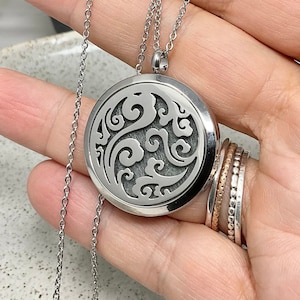 May include: A silver pendant necklace with a circular, patterned charm. The charm has an intricate, swirling design. The necklace is shown against a neutral background, highlighting the jewellery's details. The chain is delicate and complements the pendant's design.