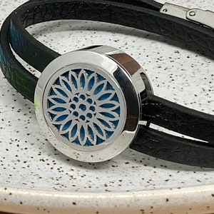 May include: Black leather bracelet with a silver aromatherapy diffuser. The diffuser features a floral design and a blue interior. Includes colourful felt pads in black, purple, blue, and green.