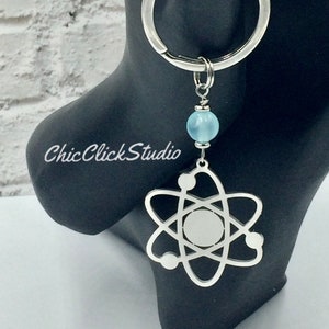 May include: Silver atom keyring with a light blue bead and a silver ring. The atom pendant features a central circle and orbiting spheres. The text "Chic Click Studio" is visible on the black surface.