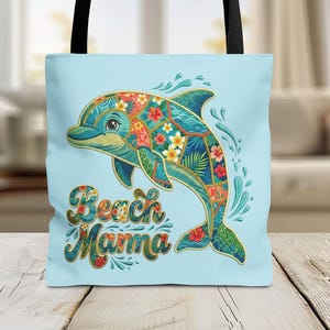 May include: Light blue tote bag featuring a colorful dolphin design with floral and tropical leaf patterns. The dolphin is outlined in gold and the text "Beach Mamma" is displayed below. The bag has black handles.