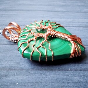 May include: A green malachite pendant with a copper wire tree design. The pendant hangs from a silver chain. The copper wire is wrapped around the stone, forming the shape of a tree.