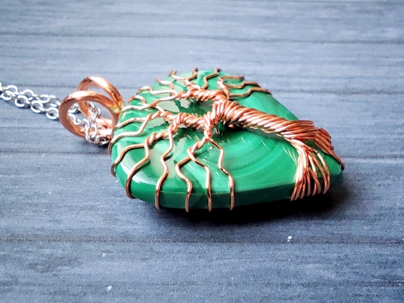 May include: A green malachite pendant with a copper wire tree design. The pendant hangs from a silver chain. The copper wire is wrapped around the stone, forming the shape of a tree.