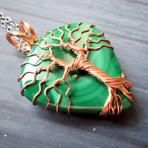 May include: A green malachite pendant with a copper wire tree design. The pendant is on a silver chain. The tree design is wrapped around the malachite stone. The pendant is a unique piece of jewelry.