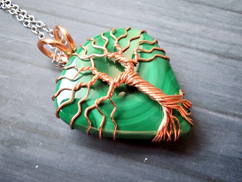May include: A green malachite pendant with a copper wire tree design. The pendant is on a silver chain. The tree design is wrapped around the malachite stone. The pendant is a unique piece of jewelry.