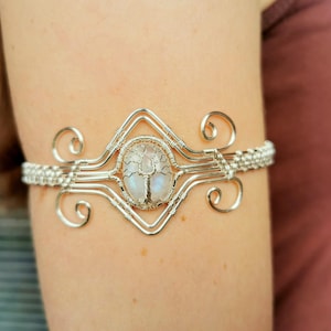 Pode incluir: Pulseira de fio de prata com um amuleto de árvore da vida de pedra da lua. A pulseira é desenhada com um padrão delicado e intrincado.