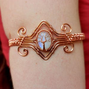 Può includere: Bracciale color rame con un design ovale centrale con un motivo ad albero della vita. Il bracciale è realizzato con filo metallico intrecciato in modo intricato con volute decorative. La pietra centrale ha un colore blu e bianco. Un gioiello unico.