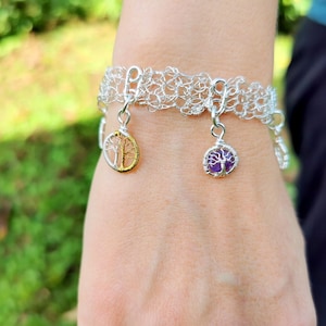 May include: A silver-toned bracelet with a woven design. The bracelet features two charms: one with a gold-colored tree design and the other with a purple stone tree design. The bracelet is worn on a wrist.