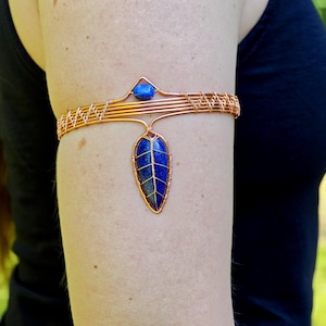 Può includere: Bracciale da braccio in rame con fascia intrecciata e pendente a forma di foglia. Il pendente è una pietra blu scuro con venature dorate. Una piccola pietra blu adorna la parte superiore del bracciale.