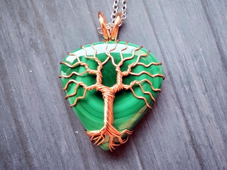 May include: A handcrafted pendant featuring a polished, teardrop-shaped malachite stone. The green stone is intricately wrapped with copper wire in a tree-of-life design, suspended from a silver chain. The pendant's dimensions are approximately 1.5 inches in length.