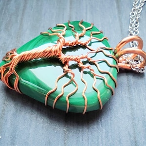 May include: A handcrafted pendant necklace featuring a polished green stone wrapped with copper wire in a tree of life design. The pendant hangs from a silver-colored chain. The copper wire has a natural, warm tone, contrasting with the vibrant green stone.