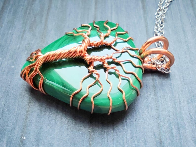 May include: A handcrafted pendant necklace featuring a polished green stone wrapped with copper wire in a tree of life design. The pendant hangs from a silver-colored chain. The copper wire has a natural, warm tone, contrasting with the vibrant green stone.