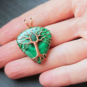 May include: A handcrafted pendant featuring a green malachite stone in a teardrop shape, intricately wrapped with copper wire to form a tree design. The pendant has a gold-colored bail for hanging.