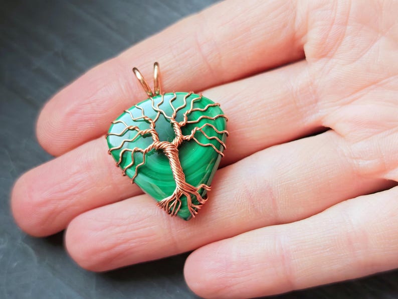 May include: A handcrafted pendant featuring a green malachite stone in a teardrop shape, intricately wrapped with copper wire to form a tree design. The pendant has a gold-colored bail for hanging.