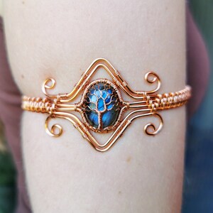Puede incluir: Brazalete de alambre de cobre con un diseño ovalado central con una piedra azul y verde, posiblemente labradorita, en un engaste de árbol de la vida de alambre. El brazalete tiene remolinos decorativos y detalles de alambre tejido.