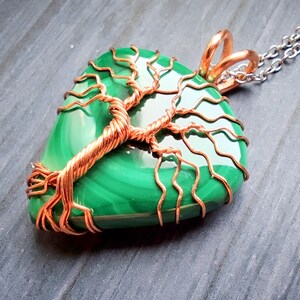 May include: A handcrafted pendant featuring a polished, green malachite stone wrapped with copper wire in a tree of life design. The pendant hangs from a silver chain. The copper wire has a natural, reddish-brown color.