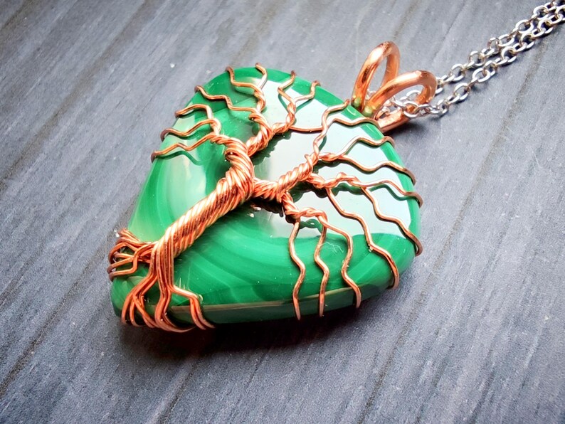 May include: A handcrafted pendant featuring a polished, green malachite stone wrapped with copper wire in a tree of life design. The pendant hangs from a silver chain. The copper wire has a natural, reddish-brown color.