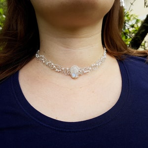 May include: A silver choker necklace with a white oval pendant. The necklace is made of small silver beads and wire.