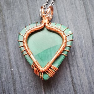 May include: A handcrafted pendant featuring a heart-shaped, green malachite stone wrapped in copper wire. The wirework forms a decorative frame around the stone, with a silver chain attached at the top. The pendant is displayed on a wooden surface.
