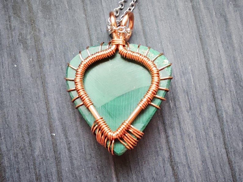 May include: A handcrafted pendant featuring a heart-shaped, green malachite stone wrapped in copper wire. The wirework forms a decorative frame around the stone, with a silver chain attached at the top. The pendant is displayed on a wooden surface.