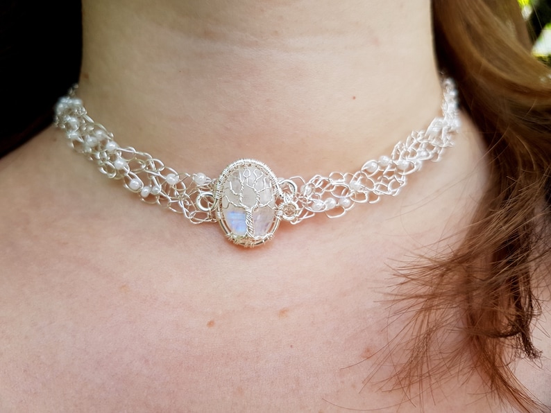 May include: A silver wire choker necklace with a silver wire tree of life pendant featuring a moonstone. The necklace is accented with small white beads.