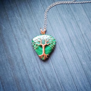 May include: A pendant necklace featuring a teardrop-shaped green stone with a copper-colored tree design. The pendant hangs from a silver-colored chain. The necklace is displayed on a blue wooden surface.