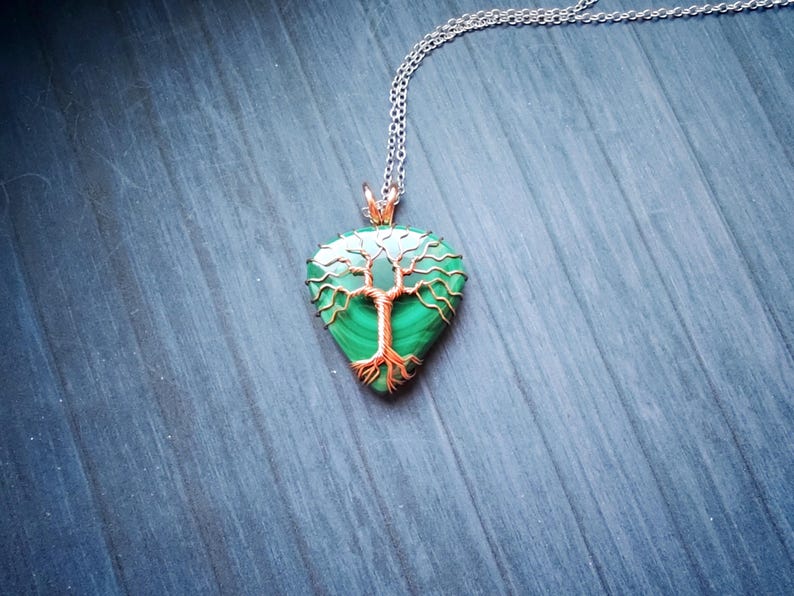 May include: A pendant necklace featuring a teardrop-shaped green stone with a copper-colored tree design. The pendant hangs from a silver-colored chain. The necklace is displayed on a blue wooden surface.