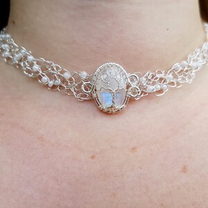 May include: A silver wire choker necklace with a white moonstone pendant featuring a tree design. The pendant is oval shaped and has a wire frame.