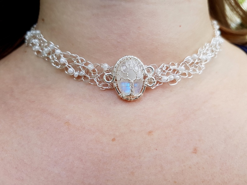 May include: A silver wire choker necklace with a white moonstone pendant featuring a tree design. The pendant is oval shaped and has a wire frame.