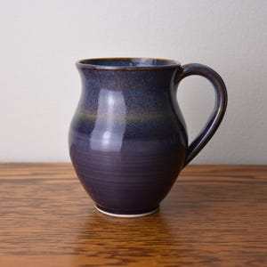 May include: A handmade ceramic mug with a dark blue and purple glaze. The mug has a rounded body, a curved handle, and a slightly flared rim. The mug is approximately 5 inches tall.