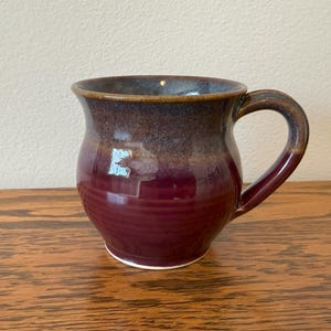May include: A handmade ceramic mug with a deep burgundy glaze that fades to a brown rim. The mug has a rounded body, a sturdy handle, and a white base. The mug is sitting on a wooden surface.