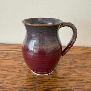 May include: A handmade ceramic mug with a deep burgundy base, transitioning to a dark purple and brown rim. The mug has a curved handle and a glossy finish, perfect for enjoying hot beverages. The mug sits on a wooden surface.