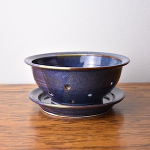 May include: A handmade ceramic bowl and saucer set. The bowl is a deep blue with brown accents and small drainage holes. The saucer is a matching dark blue. The set is displayed on a wooden surface.